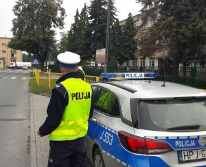 Dwóch policjantów w kamizelkach i białych czapkach stoi przy radiowozie zaparkowanym przy szkole.