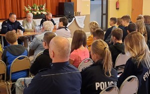 Dwóch policjantów w mundurach i mężczyzna w marynarce siedzą za stołem i rozmawiają z osobami siedzącymi naprzeciwko nich.