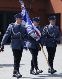 Trzech policjantów w mundurach galowych kroczy niosąc sztandar Komendy Powiatowej Policji w Namysłowie.