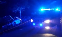 Oznakowany radiowóz policyjny stoi na drodze i ma włączone sygnały świetlne barwy niebieskiej. Obok samochód osobowy w przydrożnym rowie.