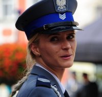 Policjantka w mundurze galowym i czapce służbowej, obrócona w prawy bok, uśmiecha się.