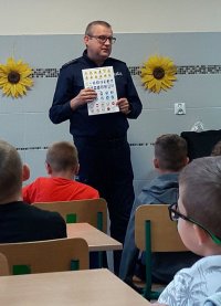 Mężczyzna w policyjnym mundurze prezentuje dzieciom znaki drogowe narysowane w książce.