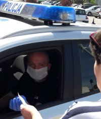 Policjant w oznakowanym radiowozie przekazuje maseczkę ochronną dla kobiety.