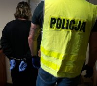 Mężczyzna w kamizelce odblaskowej z napisem policja stoi obok kobiety z założonymi kajdankami na ręce trzymane z tyłu.