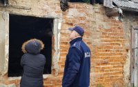 Policjant w mundurze wraz z drugą osobą w kapturze na głowie zaglądają przez dziurę w ścianie do środka budynku.