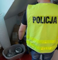 Mężczyzna w kamizelce z napisem policja stoi w mieszkaniu przy pakunkach.