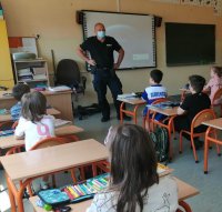 Policjant w mundurze stoi w klasie i rozmawia z dziećmi siedzącymi w ławkach szkolnych.