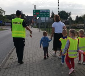 Policjantka w mundurze idzie z grupą dzieci po chodniku, wszyscy mają założone kamizelki odblaskowe.