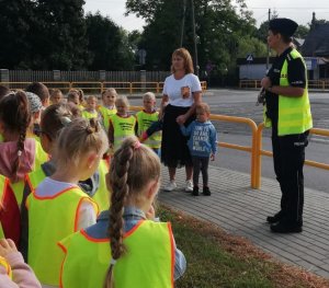 Policjantka w mundurze stoi z grupą dzieci po chodniku, wszyscy mają założone kamizelki odblaskowe.