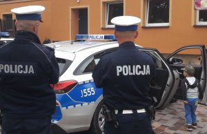 Dwóch policjantów w mundurach stoi przy radiowozie do którego wsiada dziecko.