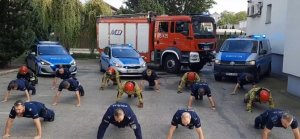 Umundurowani policjanci i strażacy robią pompki na placu, w tle radiowozy.