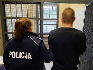 Policjantka w mundurze wprowadza do okratowanego pomieszczenia mężczyznę w dresie.