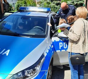 Dzieci siedzą w policyjnym radiowozie, który ma otwarte drzwi, obok stoi opiekunka i policjant w mundurze.