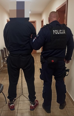 Policjant w mundurze trzyma pod rękę mężczyznę w dresie, zakutego w kajdanki zespolone.