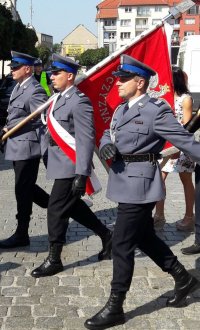 Trzech umundurowanych mężczyzn w galowe mundury policyjne kroczy krokiem defiladowym niosąc sztandar.