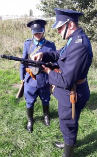 Mężczyźni przebrani w mundury policyjne z 1939 roku oglądają karabin maszynowy, trzymany przez jednego z nich.