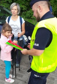 Mężczyzna w policyjnym mundurze i kamizelce odblaskowej podarowuje opaskę odblaskową dla małej dziewczynki.