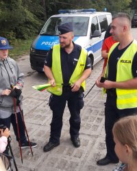 Policjanci w mundurach trzymają w ręku opaski odblaskowe i po kolei przekazują je dla ludzi zgromadzonych naprzeciwko nim.