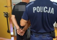 Policjant w mundurze trzyma za rękę mężczyznę z kajdankami na rękach.