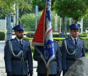 Trzech policjantów w mundurach galowych stoi na baczność, środkowy trzyma pionowo sztandar.