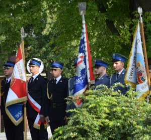 Sześciu mężczyzn w galowych mundurach strażaków i policjantów stoi w rzędzie z dwoma sztandarami.