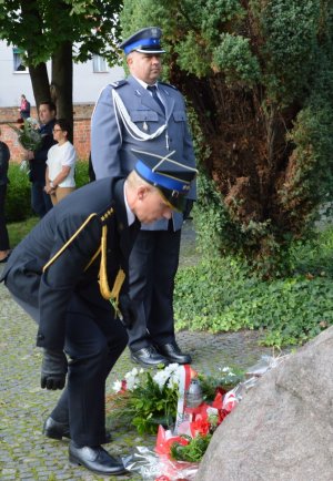 Mężczyzna w mundurze galowym straży pożarnej składa kwiaty pod obeliskiem, obok na baczność stoi policjant w mundurze galowym.