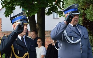 Salutuje dwóch mężczyzn w mundurach galowych straży pożarnej i policji.