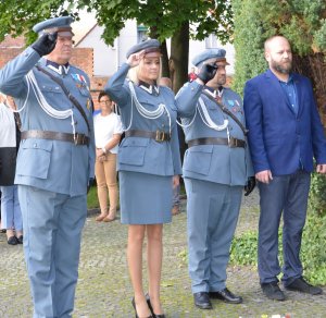 Trzy osoby w mundurach piłsudczyków oddaje honor przed obeliskiem, obok mężczyzna w garniturze.