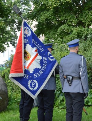 Trzech policjantów w mundurach galowych ze sztandarem stoi tyłem do aparatu.